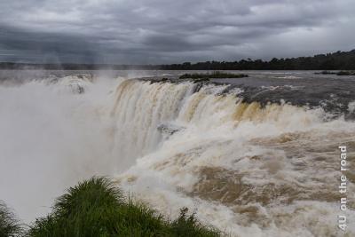Bild Iguazu - Teufelsschlucht Impression 1; es zeigt den als Teufelsschlucht benannten tiefen Kessel an den Iguazu Wasserfällen, in den von fast allen Seiten tosend Wasserfälle stürzen. Nur durch eine Schmale Öffnung kann das Wasser abfliessen. Eine permanente Gischtwolke steht in und über dem Kessel.