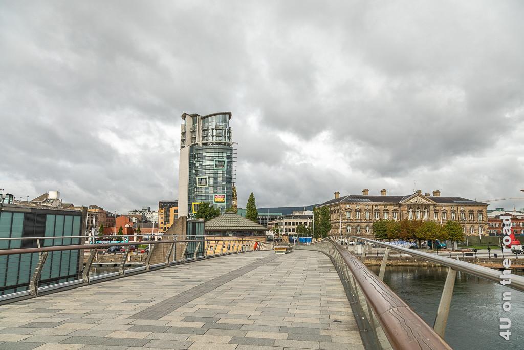 Bild Blick zurück vom Ufer des Titanic Viertels in die Stadt, Belfast zeigt die Brücke, einen lustigen Büroturm, der wie Bilderrahmen aussen hängen hat. Auf der anderen Seite der Brücke steht ein grosses Haus aus dem letzten Jahrhundert.