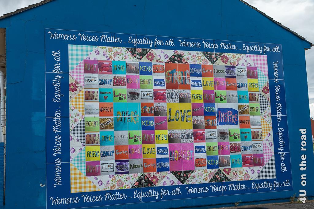 Bild Womans Quilt, Black Taxi Tour Belfast zeigt auf mittelblauer Hauswand das Bild eines riesigen, farbenprächtigen Flickenteppichs.