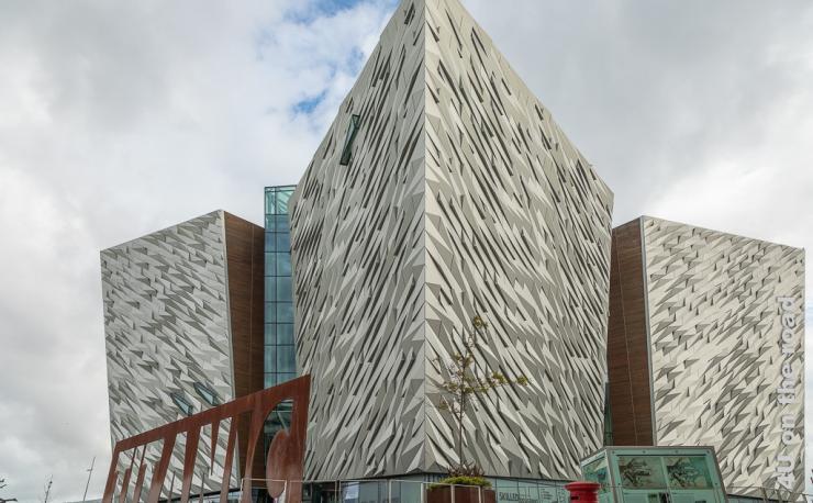 Bild Titanic Museum, Belfast zeigt das imposante Gebäude des Museums von der Seite. Man sieht drei Dreiecke mit glänzender Oberfläche in Kristallstruktur. Davor steht in rostigen Lettern Titanic. Um das Gebäude sind Bäume in ebenfalls rostigen Behältern gepflanzt.