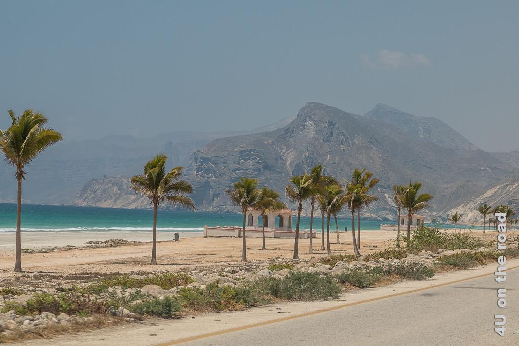 Bild Der Mughsail Beach mit Palmen und Picknickplätzen auf Höhe des Fischerdorfes zeigt den hinteren Strandabschnitt. Im Hintergrund erheben sich recht nah die hohen Klippen. Der Himmel ist von Sand gefärbt eher graublau.