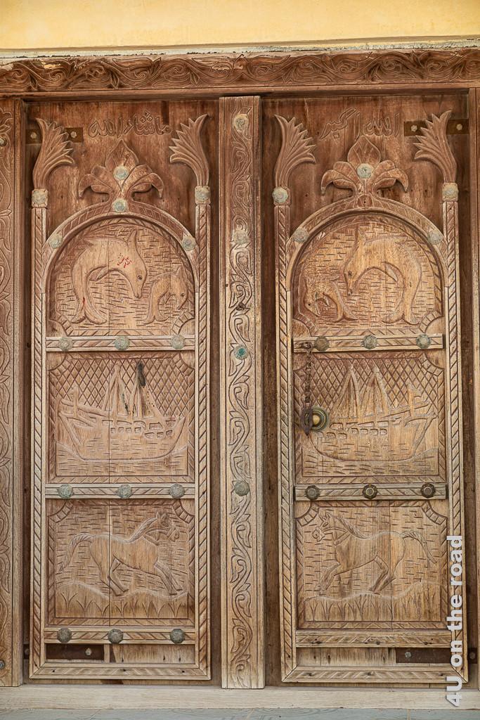 Ras al Hadd - schöne alte Holztür. Die zweiflügelige Holztür hat geschnitzte Motive: unten galopierendes Pferd, in der Mitte ein Dhau mit Segeln, oben zwei springende Delfine.