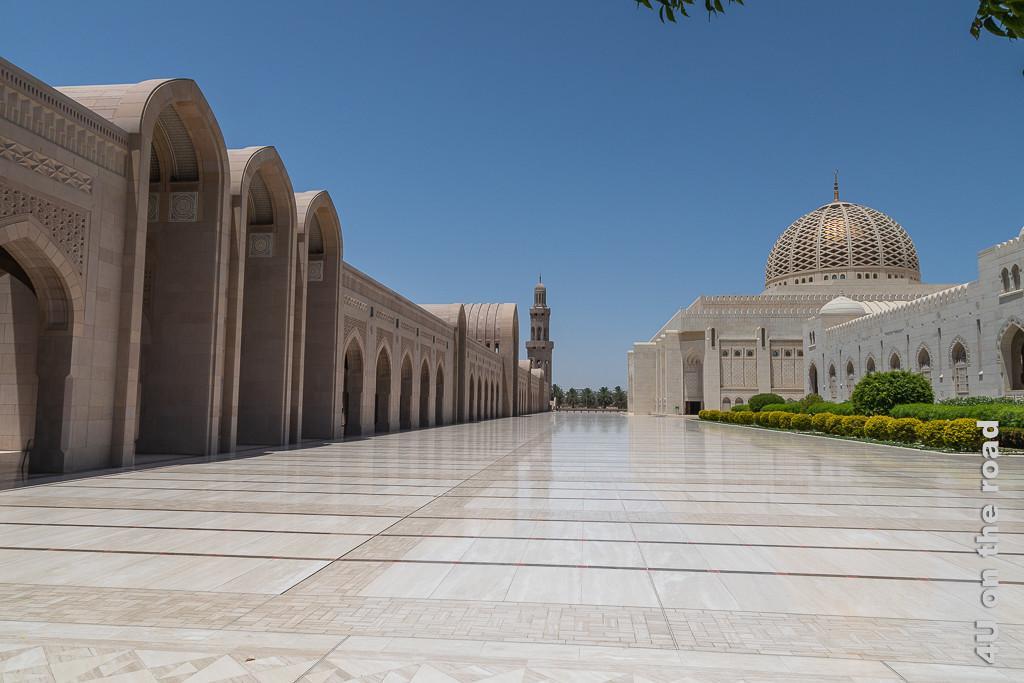 Dieses Bild vermittelt einen Eindruck der unglaublichen Grösse der Sultan Qaboos Moschee in Muscat. Rechts ein in rötlich schimmerndem Stein gehaltener Säulengang, der von einem Minarett beendet wird. Links Gebäude mit grüner Rabatte, dahinter die Moschee und zwischen den Gebäuden ein breiter Gang, der mit hellem Marmor gefliest ist.