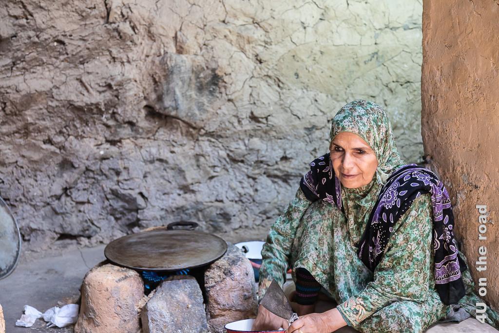 Im Museum Bait al Safah, Al Hamra - Auf dieser Eisenplatte wird das Brot gebacken. Im Bild sieht man eine ältere Frau auf einem Kissen auf dem Boden sitzend, mit den Händen in der Teigschüssel. Neben ihr bilden aufgeschichtete grosse Steine, um eine Gasflamme den Herd. Auf den Steinen liegt lose eine Metallplatte.