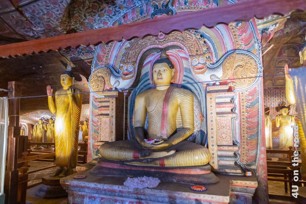 Im Vordergrund sitzt ein grosser Buddha auf einem Thron. Daneben steht ein Buddha mit erhobenen Händen. Die rechte Handfläche ist offen, die linke geballt.