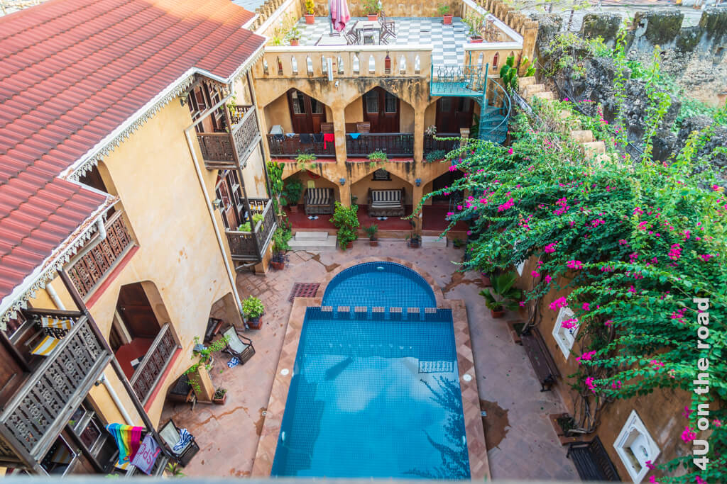 Der Innenhof des Minzingani Hotels hat einen Pool. Über die Mauer des Nachbargrundstücks wachsen Bouganvilea. Auf den anderen beiden Seiten sind Zimmer mit Balkonen.