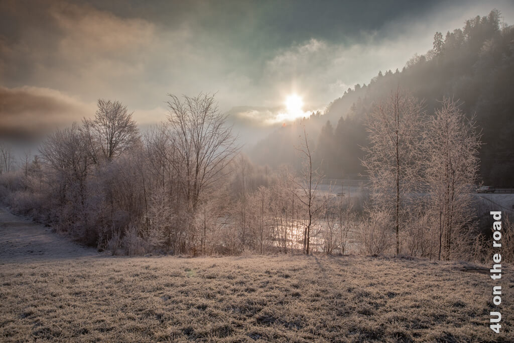 Die Sonne scheint durch die Wolken und beginnt den Nebel aufzulösen. Bäume und Gras sind voller Raureif, das Wasser des Flusses glitzert durch die Bäume. Alles ist in rötliches Licht getaucht bei dieser Momentaufnahme auf unserem Winterausflug zum Schwarzsee.