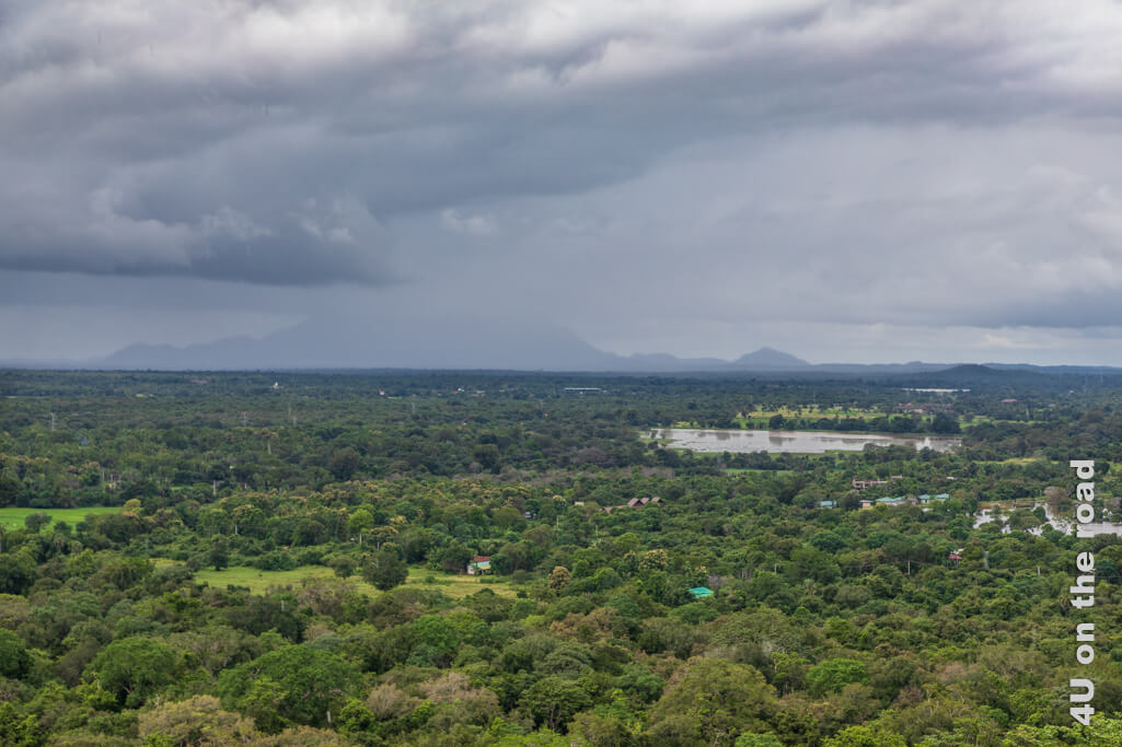 Der Blick zeigt dicke Regenwolken, die in der Ferne sich mit den Bergen im Dunst verbinden. Bis zu den Bergen gibt es eine undurchdringliche Baumfläche, nur unterbrochen durch das Glitzern von Wasseroberflächen und einigen Grasflächen. Ausblick vom Sigiriya Felsen. 
