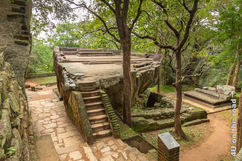 Eine Treppe führt auf die eben abgetragene Oberfläche eines einzeln in der Landschaft stehenden Felsens. Wege umrunden den Felsen und Reste von Fundamenten behaupten sich gegen die Bäume. Sigiriya Felsenfestung