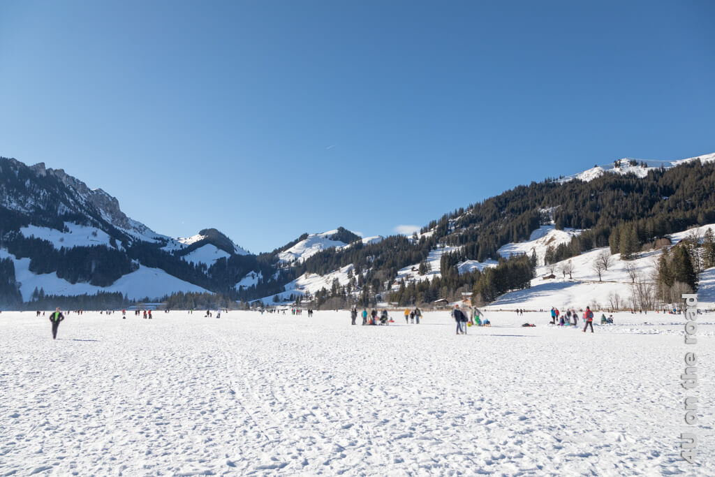 Der zugefrorene See befindet sich unter einer Schicht aus Schnee und Eiskristallen. Blauer Himmel, schneebedeckte Hügel und Berge und bunt gekleidete Menschen sind die Kulisse für den Winterausflug zum Schwarzsee.