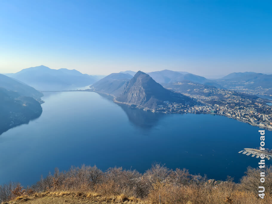 Der Luganersee mit dem Damm, einem Stück Lugano, dem Monte San Salvatore und dem langen Bergrücken nach Morcote sind im Bild zu sehen. Wandern zum Monte Brè