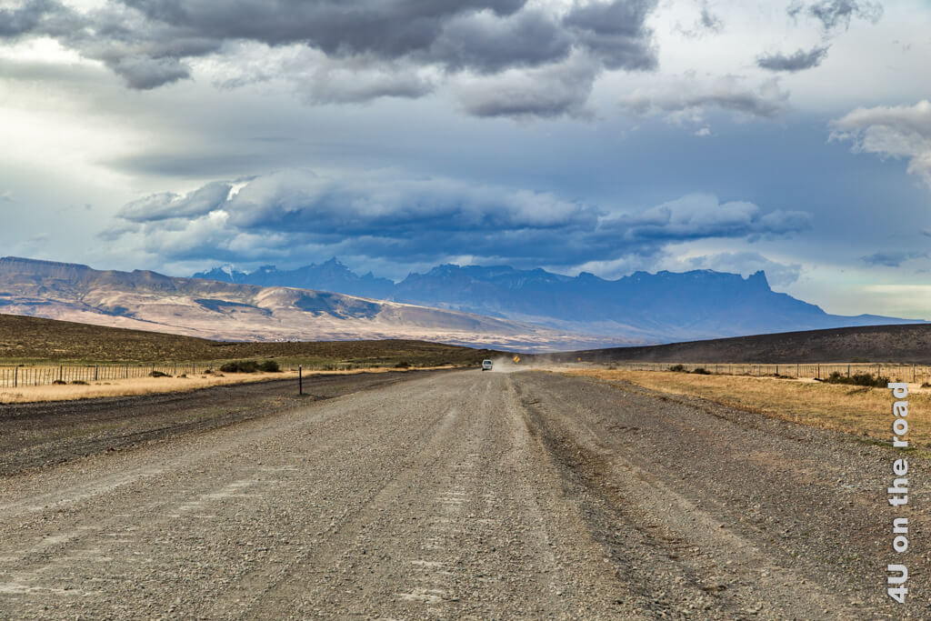 Eine breite Strasse mit Spurrillen und vielen Steinen führt zum Torres del Paine National Park, dessen Berge bereits im Hintergrund zu sehen sind. Ein Auto wirbelt Staub vor uns auf. Dunkle Wolken und sonnenbeschienene Stellen wechseln sich ab.