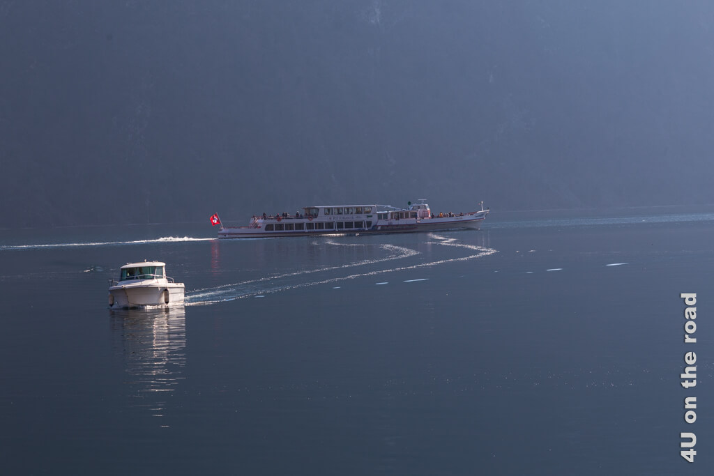 Das Schiff nach Lugano fährt aus dem Bild, während ein kleines Motorboot direkt auf den Betrachter zufährt und im spiegelglatten Wasser Spuren hinterlässt.
