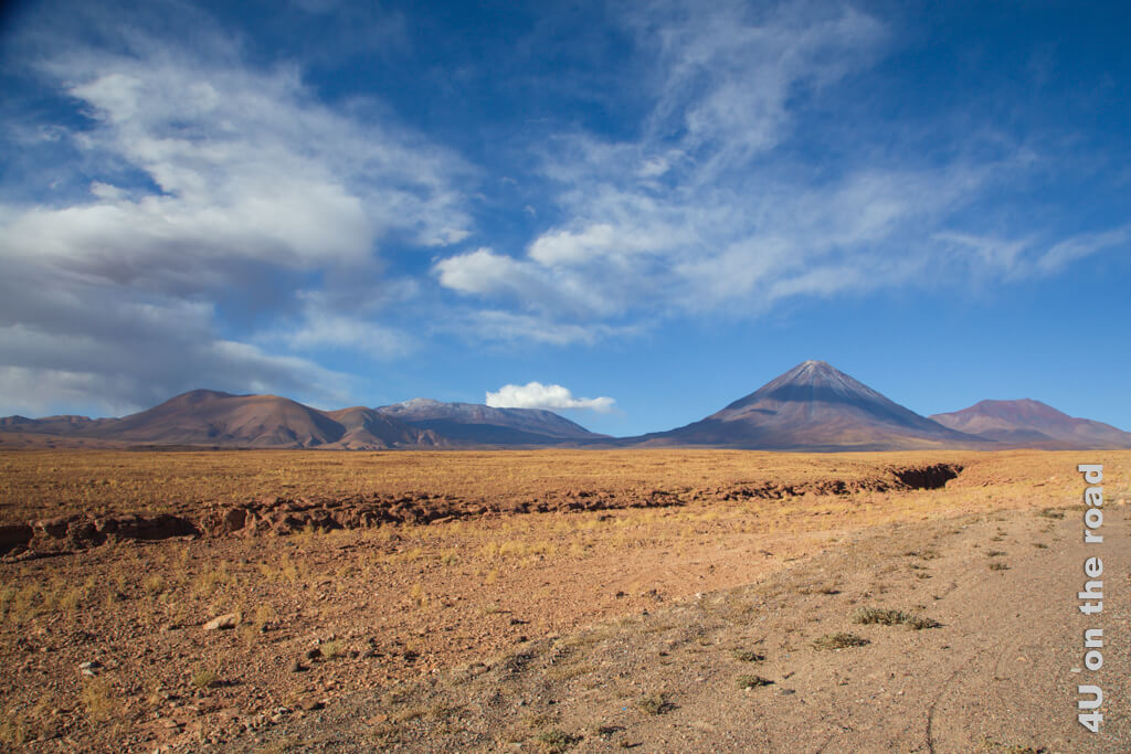 Blauer Himmel mit ein paar Wolken, ein Kegelvulkan und andere Berge, eine Gesteinswüste mit gelben Gräsern. Und absolute Einsamkeit. Sehenswürdigkeiten der Atacama Wüste