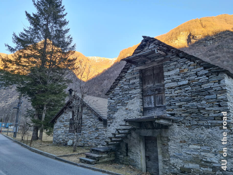 Typische Rusticos, wie die Tessiner Steinhäuser auch genannt werden, stehen an der Strasse im Schatten, während die Bergspitzen im golden Licht der untergehenden Sonne erstrahlen. Beim Wandern im Verzascatal triffst du immer wieder auf solche Häuser.