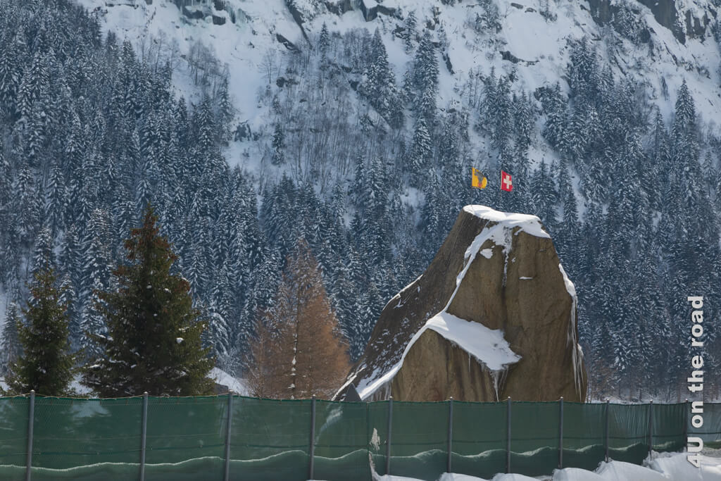 Der grosse Felsbrocken, mit dem der Teufel die Teufelsbrücke über der Schöllenenschlucht zerstören wollte, liegt direkt neben der Autobahn. Oben stecken zwei Fahnen. Die Fahne der Schweiz und die vom Kanton Uri. Auf dem Bild ist Winter und überall liegt Schnee.