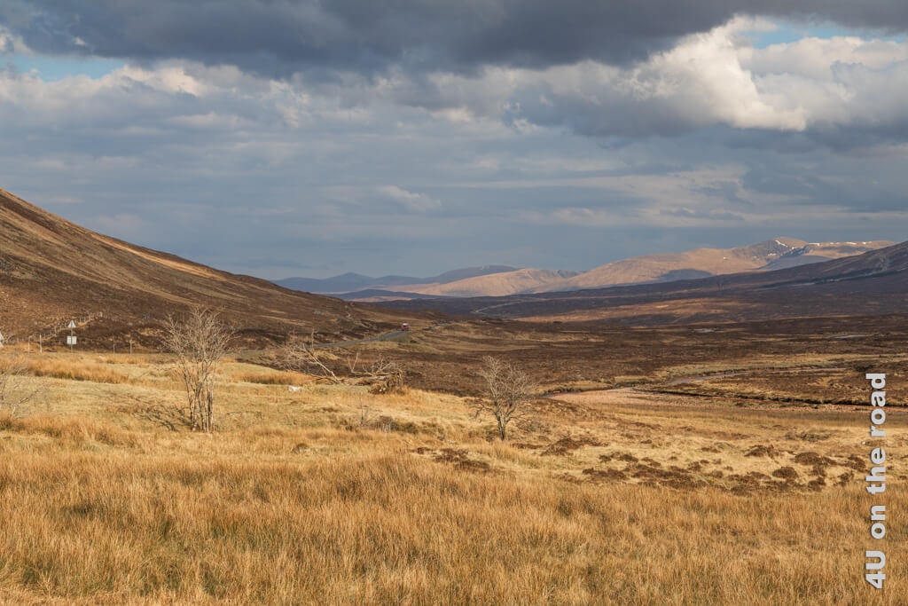 Weite und Einsamkeit. Die Strasse verschwindet am Horizont vor einer weiteren Bergkette. Der Himmel ist von dunklen Wolken überzogen, aber im Hintergrund werden die Berge von der Sonne angeleuchtet. Binsen, Gras und Heidekraut nur von einem Flusslauf und vier laublosen Bäumen unterbrochen. Das schottische Highland ist hier im Hochland beim Glen Coe Tal schmerzhaft schön. 