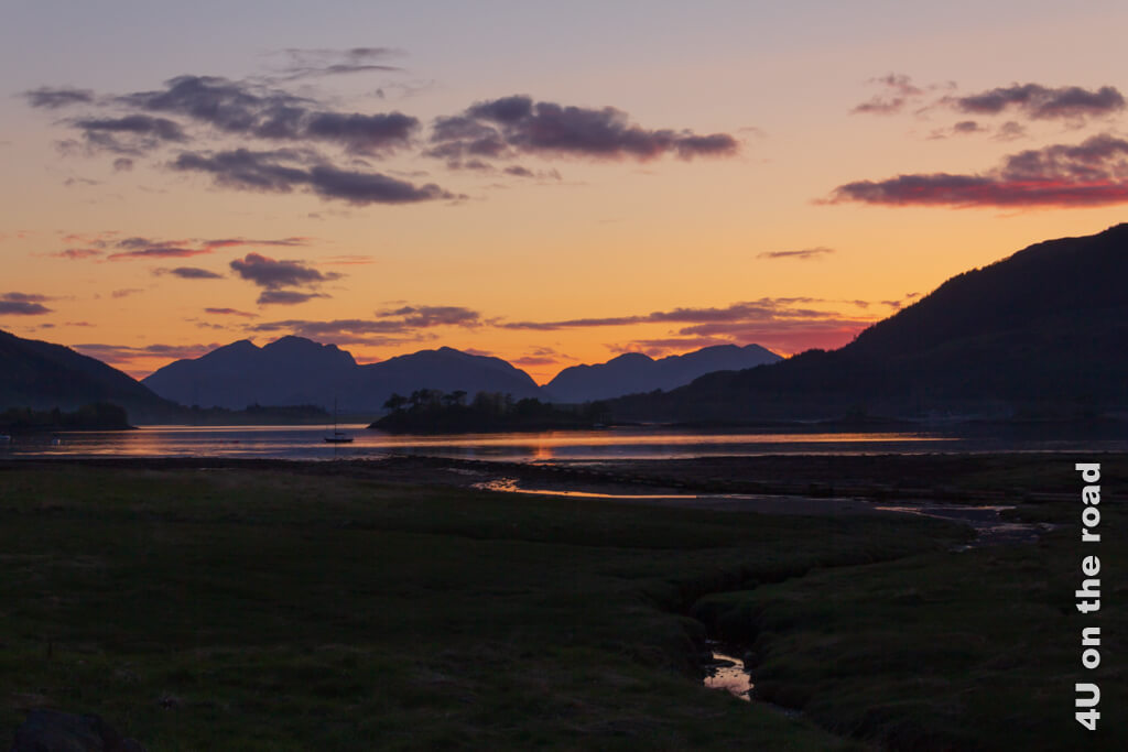 Der Himmel glüht orange über Loch Leven und die Zacken der dunklen Berge ragen in den Himmel. Das Licht der untergehenden Sonne spiegelt sich im See und dem Flüsschen. Ein Boot und eine Insel machen das Bild von Glencoe aus gesehen perfekt. So romantisch können die schottischen Highlands aussehen.