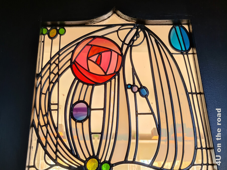 Ein Bleiglasfenster mit geraden Linien, der Mackintosh Rose in Rottönen und farbigen Tropfen in unterschiedlichen Farben.