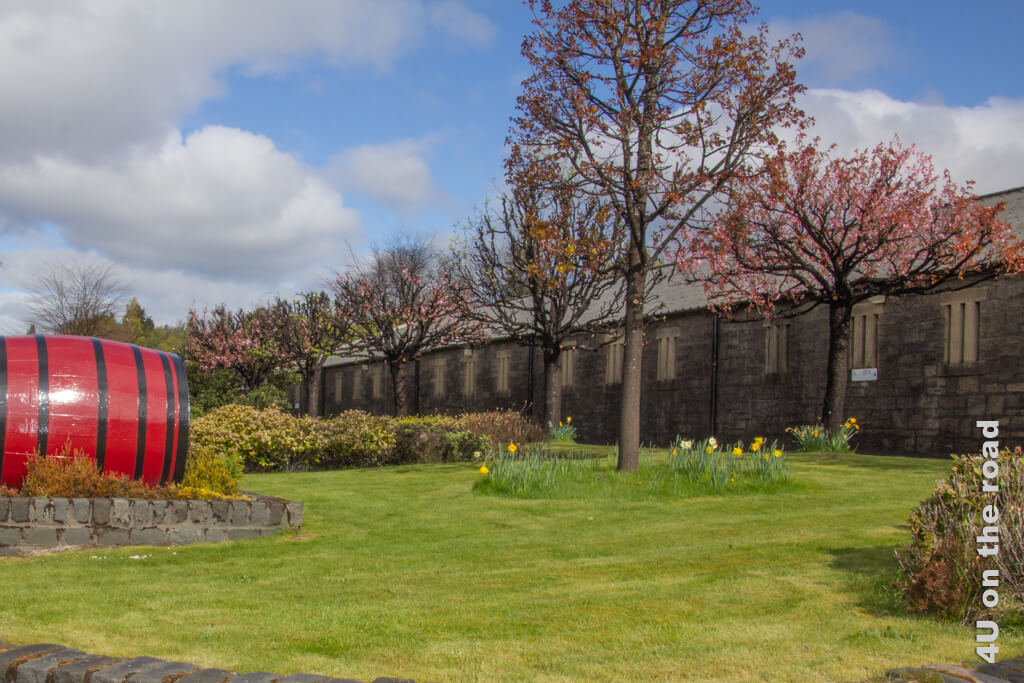 Hinter einer Wiese mit Blumenrabatten rund um mehrere Bäume erstreckt sich langgezogen das Lagerhaus der Blair Athol Destillerie. Im Vordergrund links liegt ein grosses, rotes, altes Whisky Fass.