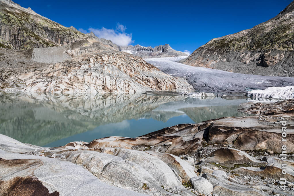 Im Bild zu sehen ist der Rhonegletscher, auch Furkapass Gletscher genannt und sein Gletschersee mit schönen Spiegelungen der umliegenden Berge, mit sehr charakteristischen Steinmustern, da die Steine an Bruchkanten sehr viel dunkler sind. Die hässliche Abdeckung über der Eisgrotte des Rhone Gletschers ist auch sichtbar. - Gletscher Schweiz