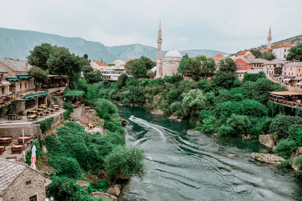 Restaurants zu beiden Seiten des Flusses warten in Mostar auf Gäste. Spitz ragen zwei Minarette in den Himmel.
