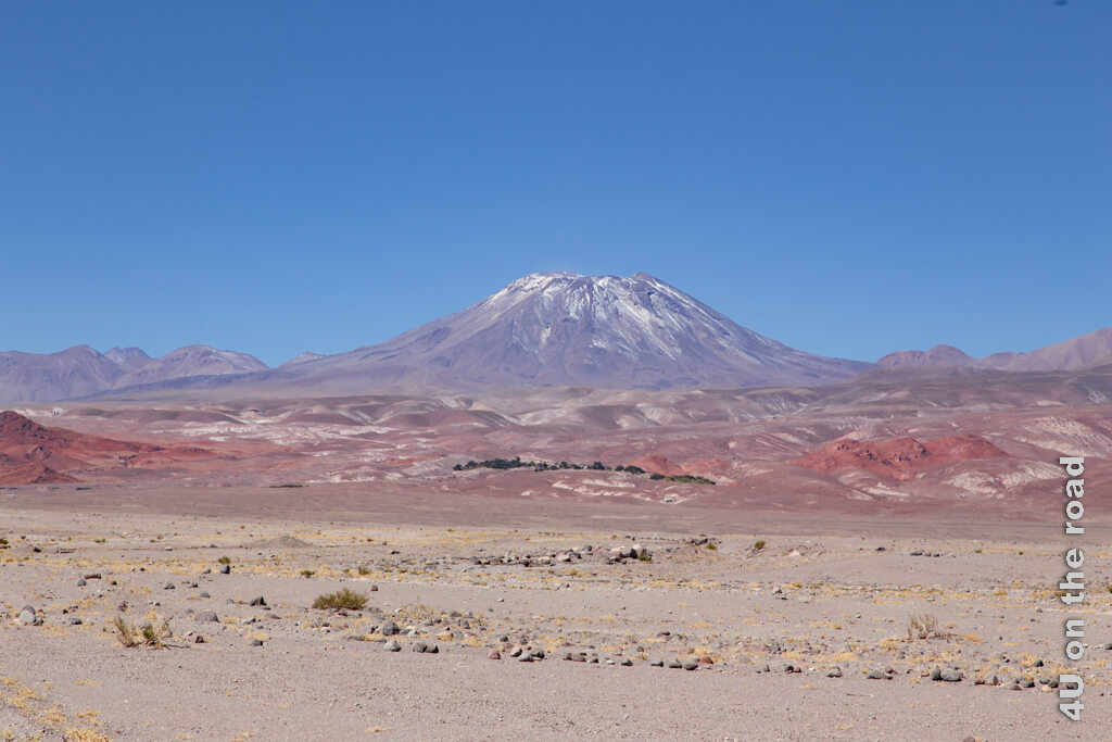 Rote, violette und weise Hügel ziehen den Blick auf einen hohen Vulkan mit Schnee auf den Gipfeln. 