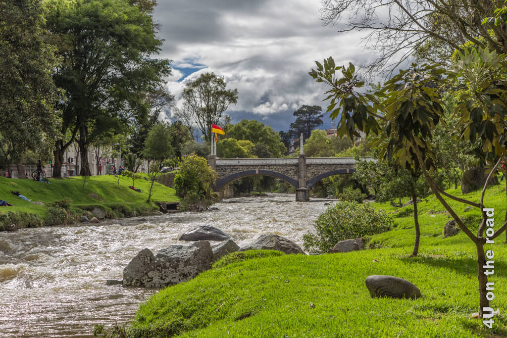Der Fluss Tomebamba fliesst durch grüne Wiesen mit schattenspendenden Bäumen. Eine Brücke führt über den Fluss in die Altstadt von Cuenca.