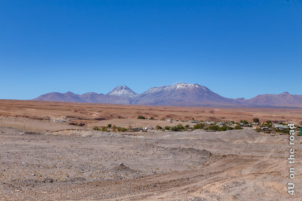 Von San Pedro de Atacama kommend, fahren wir durch Toconao. Hier sieht man die Ausläufer des Ortes in die Wüste und die Schäden der Wassererosion. Zwischen den Häuschen gibt es viel Grün, im Hintergrund erheben sich die schneebedeckten Vulkane.