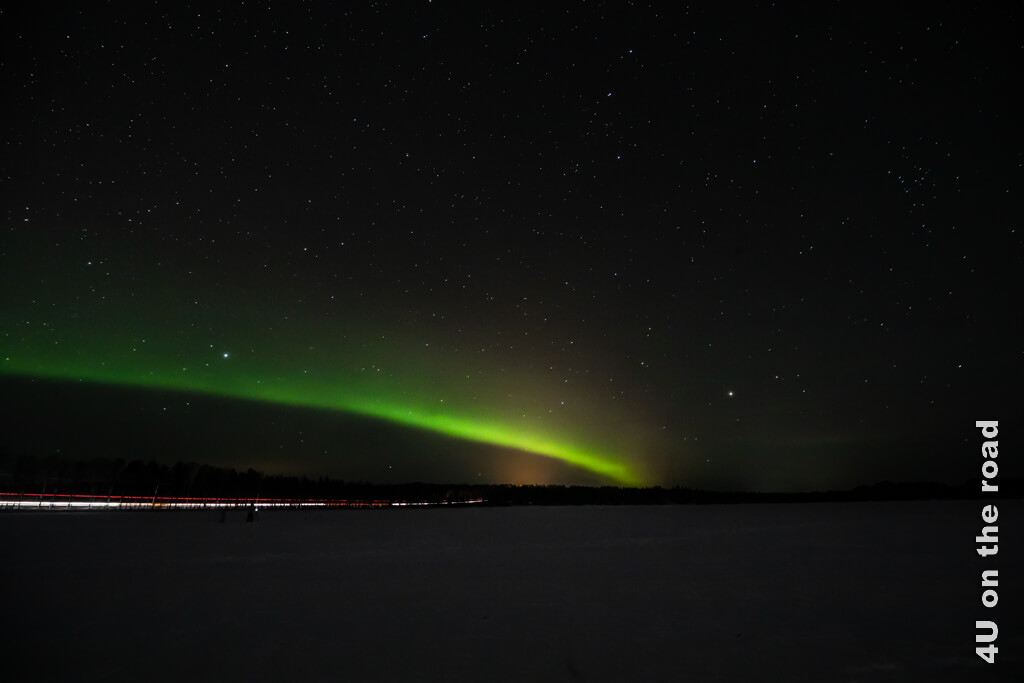 Die Strasse zum See ist noch ziemlich befahren, sodass sich rote und weisse Leuchtspuren unten durch das Bild ziehen. Die Aurora Borealis erscheint an diesem Abend erst einmal als Band in Rovaniemi.