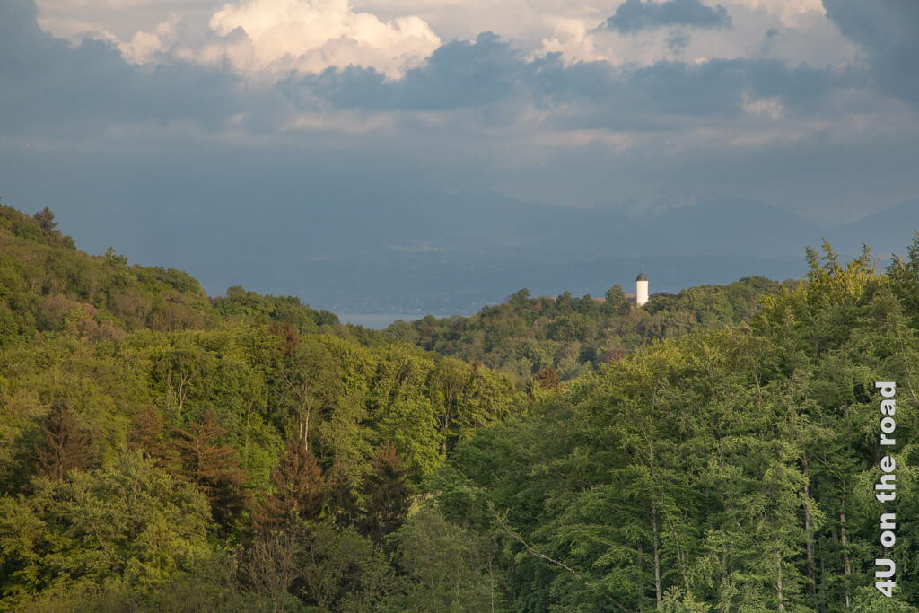 Der weisse Turm des mittelalterlichen Städtchens Aubonne im Licht der untergehenden Sonne. Der Genfersee und die Berge dahinter sind nur schemenhaft zu erkennen. Aubonne ist auch eine Sehenswürdigkeit in der Genferseeregion, die nicht nur im Frühling einen Besuch wert ist.