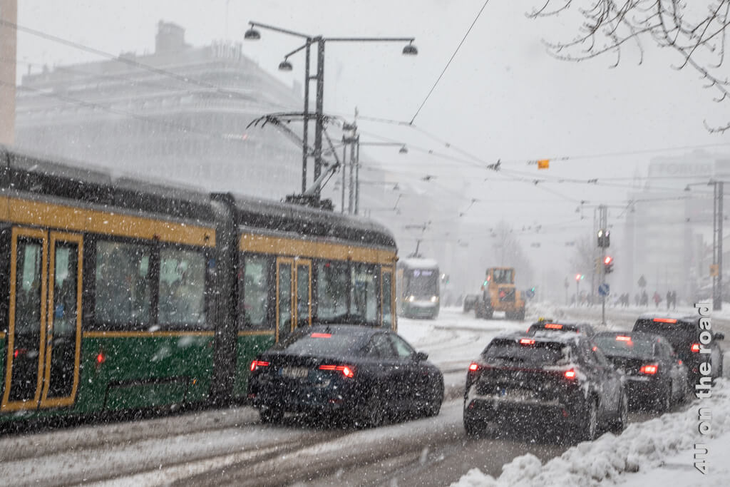 Stadtansicht von Helsinki bei starkem Schneefall mit Strassenbahnen, Räumfahrzeug und Autos, die an einer roten Ampel warten.