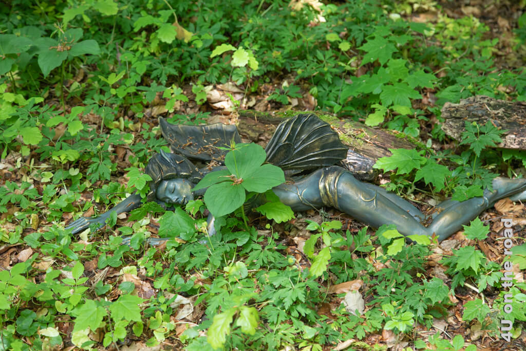 Die Bronze einer schlafenden Fee liegt auf dem Waldboden und wird vom frischen Grün der Blätter eingerahmt. Chauteau de Vullierens - Sehenswürdigkeit im Frühling Genferseeregion