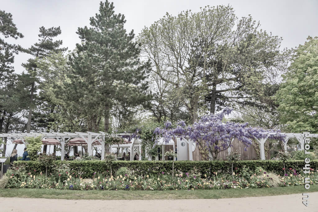 Romantisch unter hohen Bäumen von einer weissen Pergola umgeben, die von einer Glyzinie berankt wird, ist der Platz im Café in Dior's Garten in Granville.