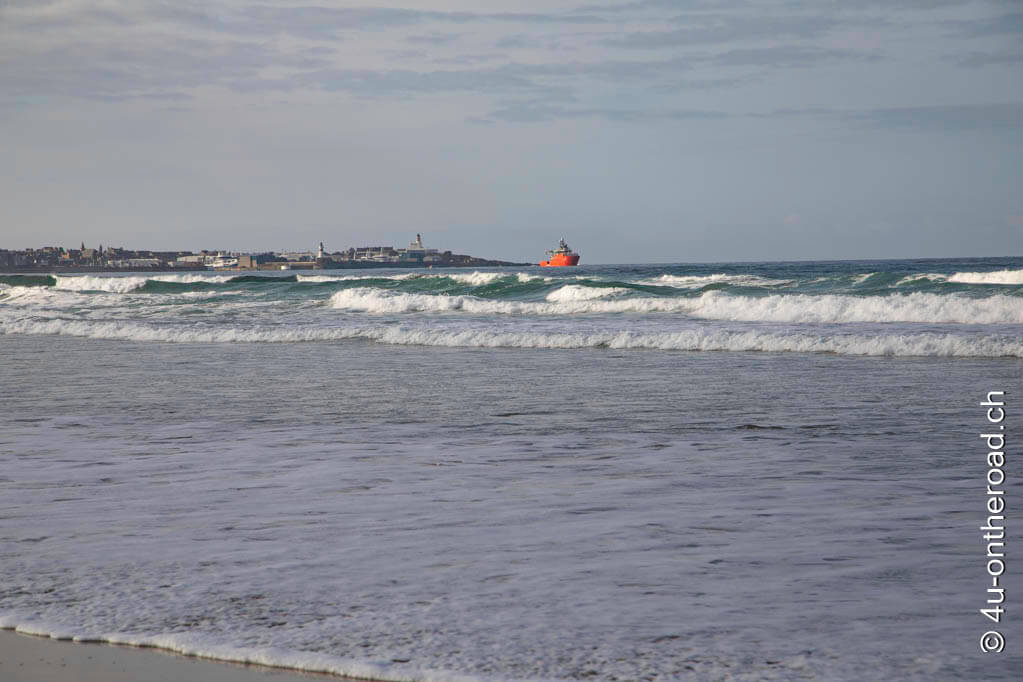 Fraserburgh ist in der Ferne zu sehen. Im Meer voller Wellen fährt ein grosses rotes Schiff raus ins offene Meer. 