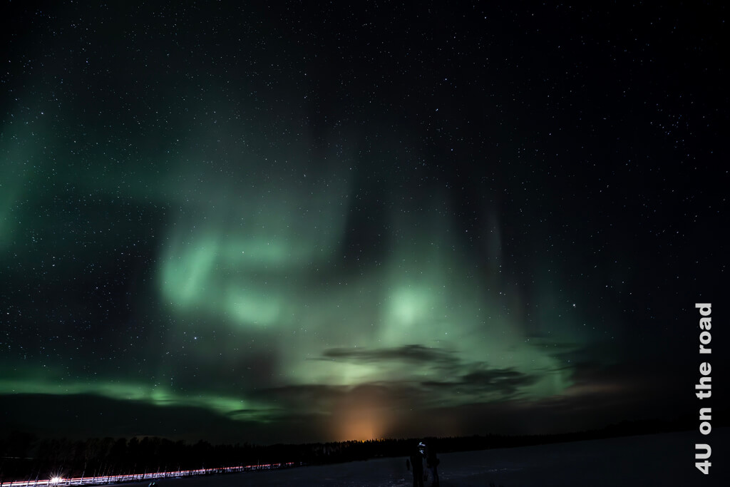 Grüne Polarlichter tanzen über den Sternenhimmel über dem orangen Lichtschein eines Ortes. Polarlichter im Jahresrückblick 2023